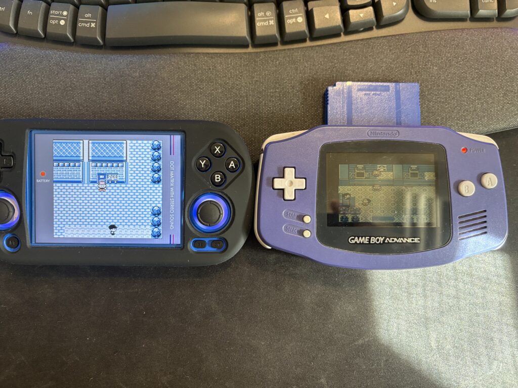 Due console portatili affiancate su una scrivania scura, sotto una tastiera da computer: a sinistra un dispositivo moderno nero (emulatore o console custom) che mostra un gioco Pokémon monocromatico in 4:3, a destra un Game Boy Advance originale viola con inserita una cartuccia blu, con lo schermo acceso che mostra un gioco.